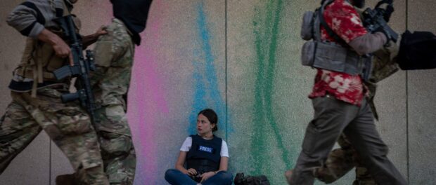 Press journalist sits on ground while soldiers march around her
